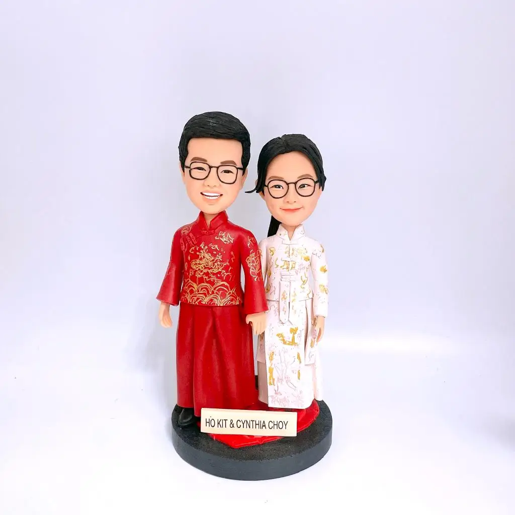 客製化Q版情侶/夫妻公仔 中式婚禮造型 (傳統裙褂/馬褂風格) 男士身穿紅色中式禮服 (飾有金色龍紋刺繡圖案) 女士身穿淡粉/白色改良式旗袍 (飾有精緻金色花紋) 雙方特徵：皆配戴黑框眼鏡、面帶幸福微笑 動作：手牽手甜蜜站姿 黑色圓形底座 客製化英文姓名牌 (印有 "HO KIT & CYNTHIA CHOY") 手工軟陶/黏土捏製 結婚禮物、婚禮蛋糕裝飾 (Cake Topper)、週年紀念、訂婚禮物