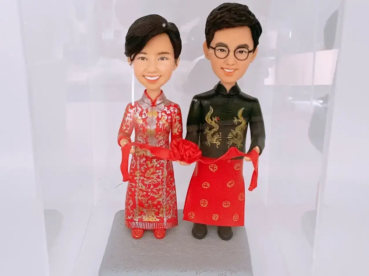 一對Q版新人穿著中式禮服牽紅綢而立：新娘著紅金秀禾服、刺繡繁複；新郎穿黑金龍紋長衫配大紅馬褂裙與圓框眼鏡，兩人微笑站在灰色底座上。