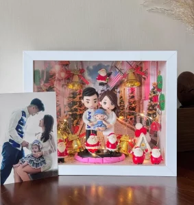 白色相框中的聖誕家庭場景：爸爸媽媽抱著嬰兒的Q版公仔站在中央，背景是聖誕燈飾街道與金色鐘、聖誕樹；前景擺有多個聖誕老人與禮物裝飾，燈串環繞增添暖光。