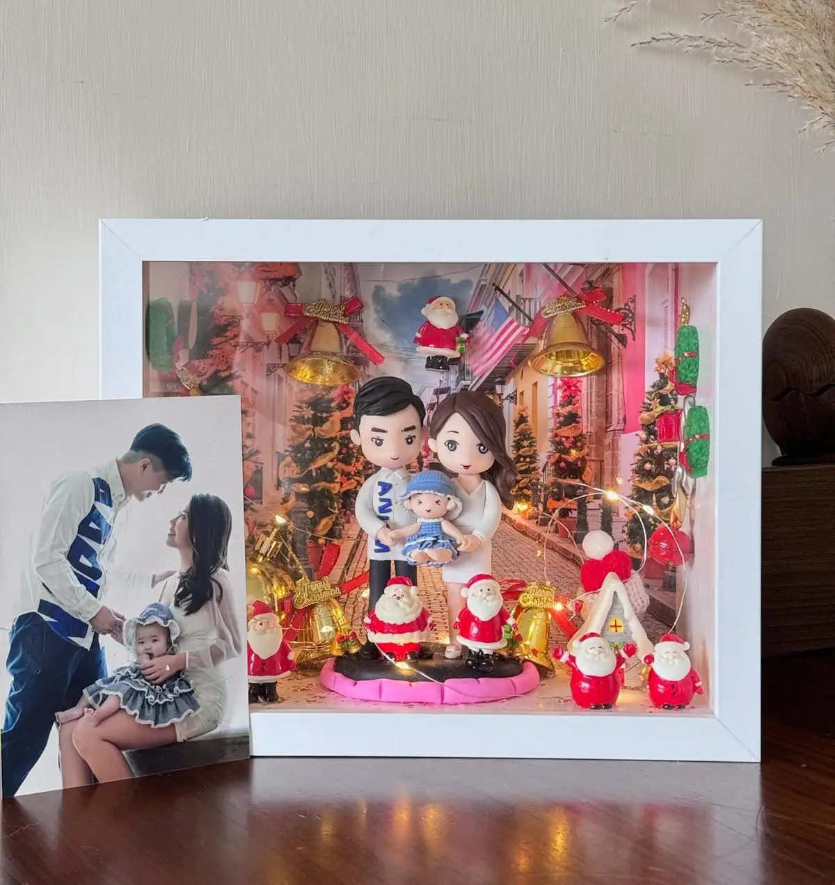白色相框中的聖誕家庭場景：爸爸媽媽抱著嬰兒的Q版公仔站在中央，背景是聖誕燈飾街道與金色鐘、聖誕樹；前景擺有多個聖誕老人與禮物裝飾，燈串環繞增添暖光。