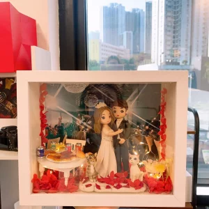 白色相框中的婚禮立體場景：Q版新娘穿白色婚紗、新郎穿黑色禮服相擁站立；腳邊有兩隻貓咪公仔與紅色花瓣。左側小桌擺放披薩、麵包與汽水罐，背景為宴會廳燈光，並以暖光燈串點綴。