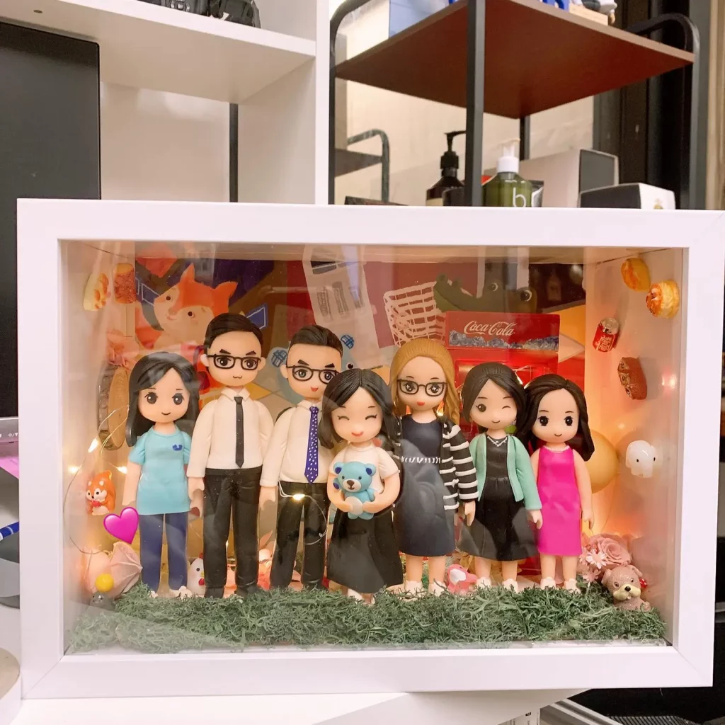 白色相框中的立體家庭場景：七位Q版人物公仔站在草地上並排合照，包含穿襯衫打領帶的兩位男性、抱著藍色小熊的女孩、戴眼鏡與穿條紋外套的女性等。背板貼滿可樂、票根、食物與寵物等元素，並有暖色燈串點綴。