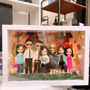 白色相框中的立體家庭場景：七位Q版人物公仔站在草地上並排合照，包含穿襯衫打領帶的兩位男性、抱著藍色小熊的女孩、戴眼鏡與穿條紋外套的女性等。背板貼滿可樂、票根、食物與寵物等元素，並有暖色燈串點綴。