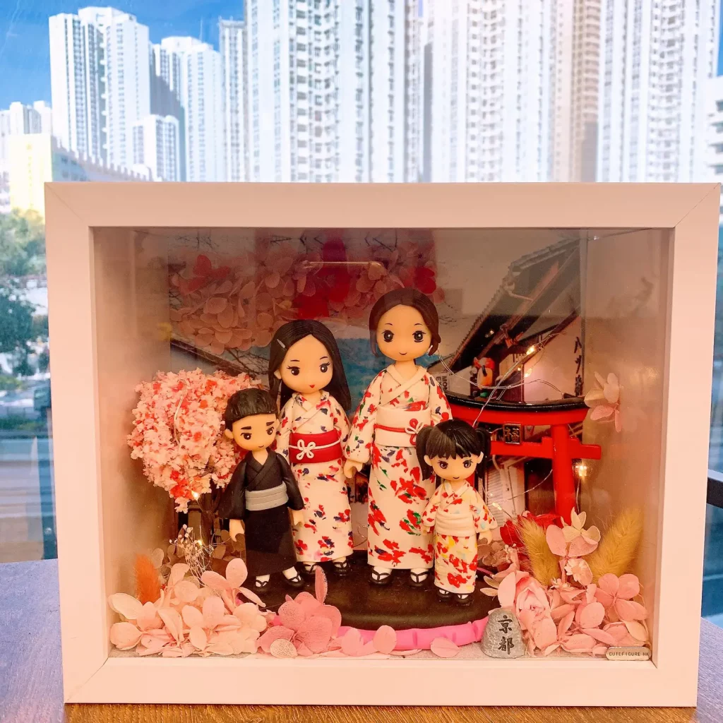 白色相框中的日式家庭場景:四位Q版公仔穿著和服,兩位大人與兩位小朋友站在櫻花樹與紅色鳥居前,前景以粉色花材與小石碑點綴,背景有日式街景與暖光燈串。