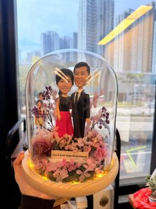 客製化情侶/週年紀念公仔玻璃盅 穿著黑色西裝男士、黑色上衣配桃紅長裙女士 圓頂透明玻璃罩 (防塵罩) 原木色圓形底座 底部裝飾粉紫色系永生花/乾燥花 (繡球花、滿天星等) 內置暖黃色LED燈串 (夜燈功能) 前方置有客製化文字小立牌 (似週年紀念祝賀語) Q版擬真/寫實風格 手工軟陶/黏土捏製 結婚週年禮物、生日驚喜、居家擺飾