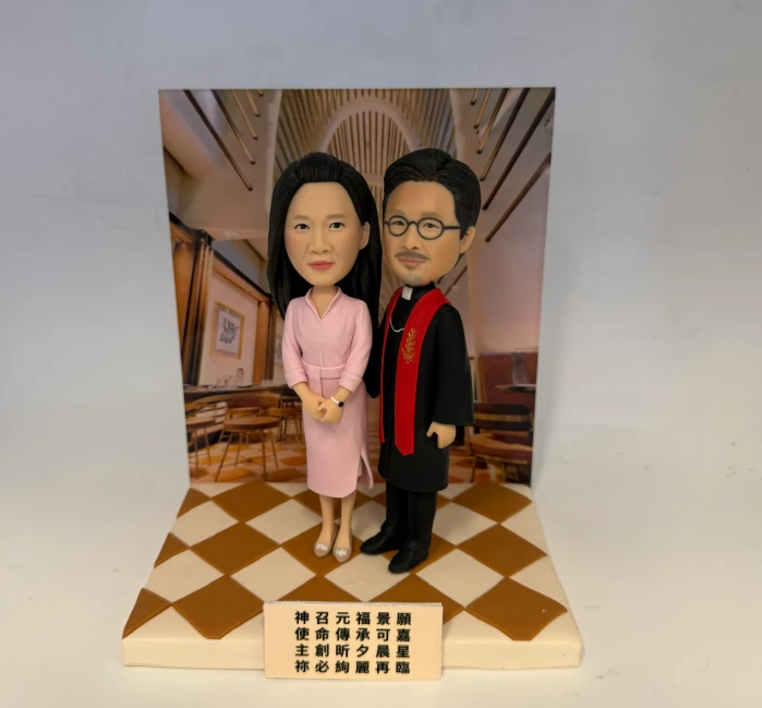教堂室內背景前的兩人公仔：女子穿粉色套裝並握手站立，男子著黑色牧師袍配紅色披帶與眼鏡，腳下是米白與棕色棋盤格地台，前方立有中文祝福小牌。
