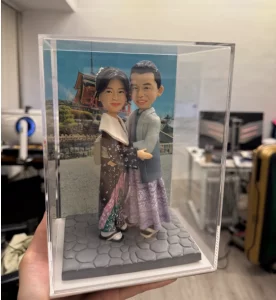 透明展示盒內的和風情侶公仔：女子穿深棕與紫綠相間的和服配披肩與木屐，男子著淡灰藍和服外搭馬甲風上衣與草履，兩人相擁站在石板路底座上，背板是日本寺院景色。