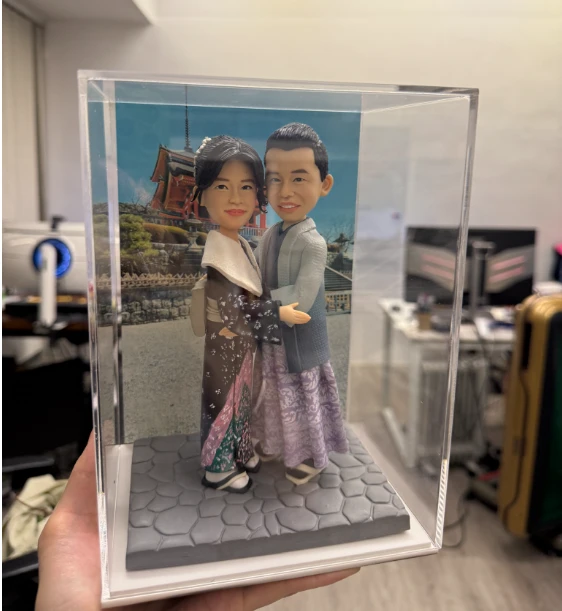 透明展示盒內的和風情侶公仔:女子穿深棕與紫綠相間的和服配披肩與木屐,男子著淡灰藍和服外搭馬甲風上衣與草履,兩人相擁站在石板路底座上,背板是日本寺院景色。