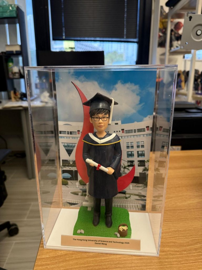 男畢業生主題人像公仔穿深藍學袍戴學士帽，手持文憑站在草地底座與校園背景前，透明展示盒內附金色名牌