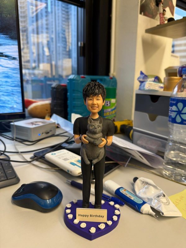 男子公仔抱著灰色貓咪站在紫色愛心底座，上緣環繞白色小玫瑰並配有「Happy Birthday」金色銘牌，生日寵物主題客製