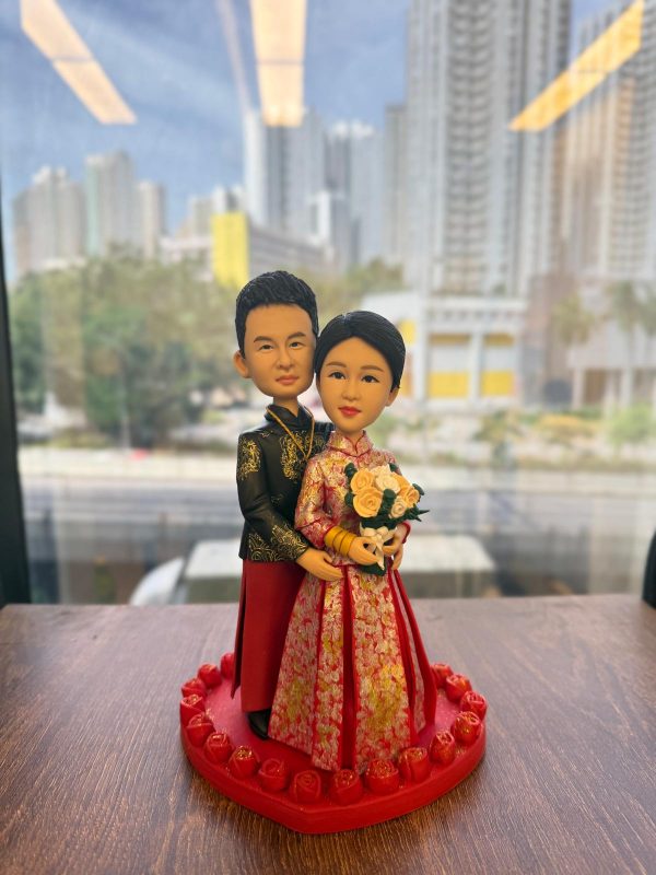 中式婚禮主題公仔：新娘穿紅金龍鳳褂手捧米白玫瑰花束，佩戴金手鐲；新郎著黑金繡紋長衫與紅裙褂，自後環抱新娘。兩人站在紅色愛心底座，周圈環繞立體玫瑰。