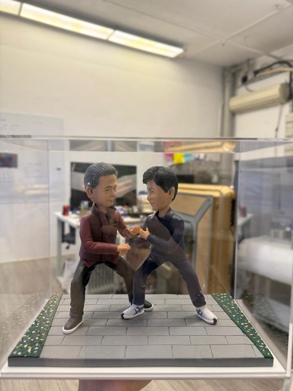 透明展示盒內的雙人公仔：兩名男子在灰色磚道地台上對練站樁交手，左側穿酒紅外套黑褲，右側著深灰運動套裝與白色武術鞋，地台邊緣有草皮與碎花點綴。