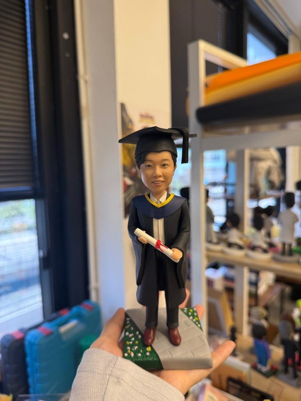 男畢業生人像公仔穿黑色學袍與方帽，手持紅綁帶文憑，站在灰色步道與綠地小花底座上，典禮感十足