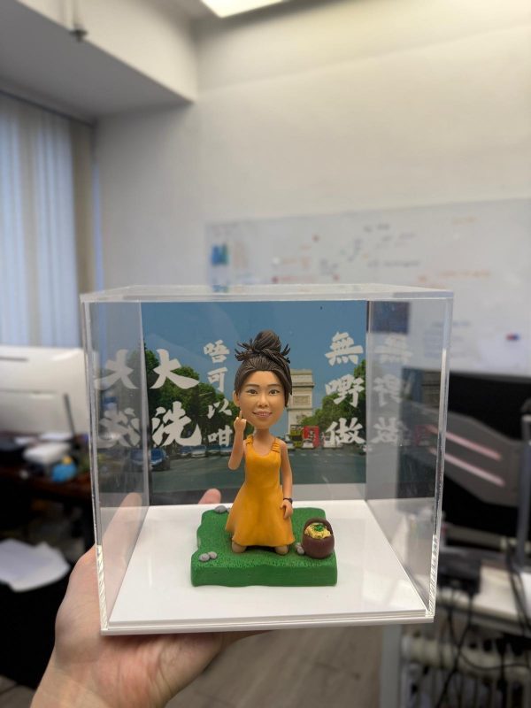 透明展示盒內的人像公仔：綁丸子頭、穿橙黃色洋裝的女孩跪坐在綠色草地地台上，右手做手勢，旁邊有小野餐籃；背板為藍天綠樹與凱旋門街景，覆有白色字樣。