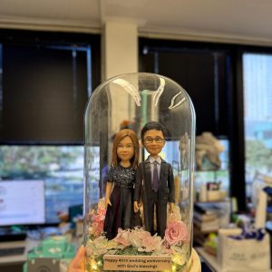 40 週年婚禮紀念雙人公仔以玻璃罩與木座呈現典雅儀式感，底部鋪滿粉色花材與暖燈，搭配客製金句銘牌。可自訂姓名、年份、服裝與祝福文字，亦能更換花色與燈光模式；精緻包裝、安全配送，讓相守不渝的愛被長久珍藏與展示。