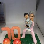 長壽主題公仔擺設：銀髮長輩穿粉色套裝抱著微笑孫子與藍色玩偶熊，綠草地台上有大型「70」與兩顆長壽桃，前方金色銘牌寫上祝福與日期。