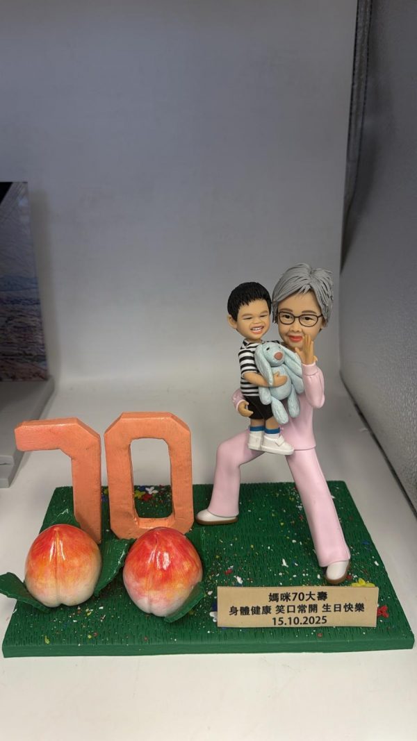 長壽主題公仔擺設：銀髮長輩穿粉色套裝抱著微笑孫子與藍色玩偶熊，綠草地台上有大型「70」與兩顆長壽桃，前方金色銘牌寫上祝福與日期。