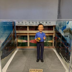 透明展示盒內的人像公仔:身穿藍色制服的男子手抱黃藍排球,站在灰色地台上;背板為「香港仔消防局暨救護站」建築外觀,前方金色銘牌刻有祝福語與編號。