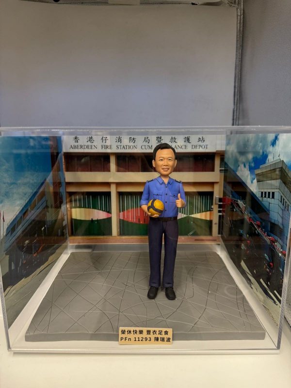 透明展示盒內的人像公仔：身穿藍色制服的男子手抱黃藍排球，站在灰色地台上；背板為「香港仔消防局暨救護站」建築外觀，前方金色銘牌刻有祝福語與編號。