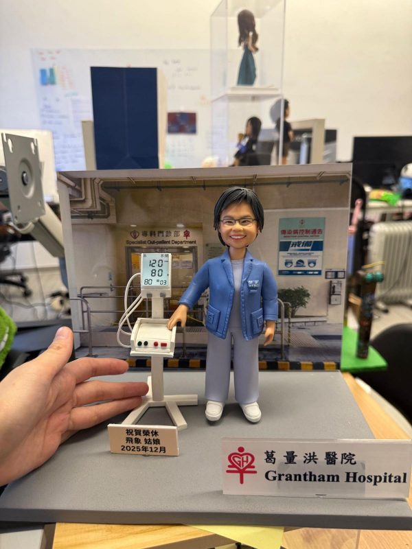 醫護主題人像公仔穿藍色外套與醫護服，站在專科門診背板前，旁邊有血壓監測儀器模型與祝賀銘牌，前方擺放「Grantham Hospital」標牌
