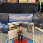 透明展示盒中的攝影主題公仔：短髮戴眼鏡的人物手持相機站在紅色愛心地墊上，背後三折屏風掛有風景照與書法字畫；地台鋪世界地圖，前方銘牌寫「Tracy 海闊天空」。