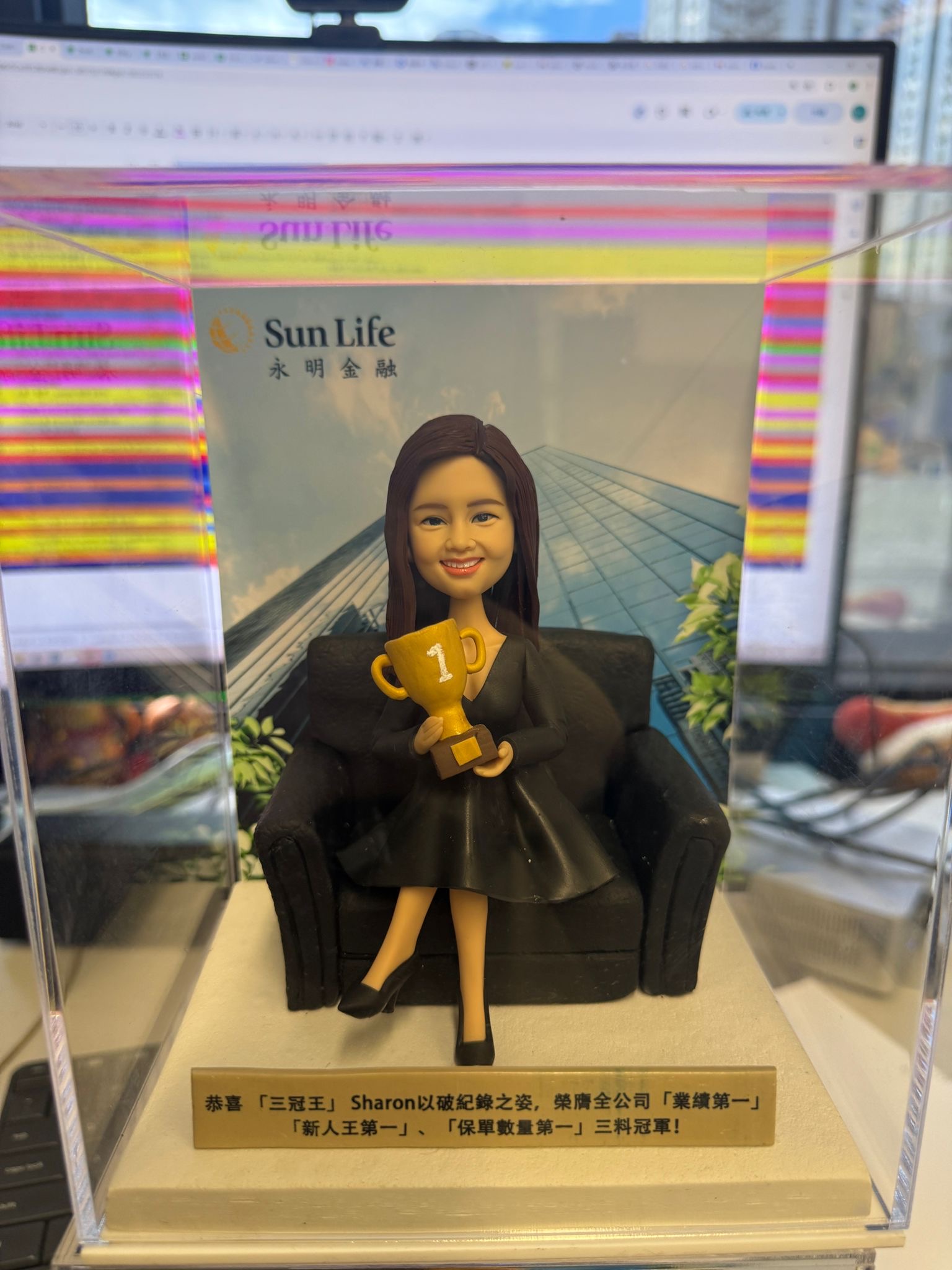 穿黑色套裝的女業務員人像公仔坐在黑色梳化上，手捧金色第一名獎盃，背後是公司大樓背景與 Sun Life 標誌