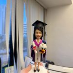 手掌托著的畢業生公仔：長髮女生戴黑色學士帽、穿紫粉鑲邊的學袍，一手拿綁紅帶的卷軸，另一手抱著可愛鴨子玩偶，站在白色圓形底座上。