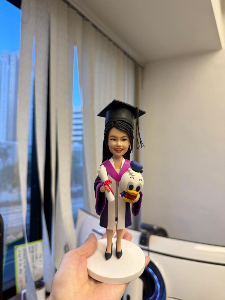 手掌托著的畢業生公仔：長髮女生戴黑色學士帽、穿紫粉鑲邊的學袍，一手拿綁紅帶的卷軸，另一手抱著可愛鴨子玩偶，站在白色圓形底座上。