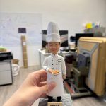 手持展示的主廚公仔：戴高高的白色廚師帽、穿白色雙排扣主廚外套與長圍裙，端著一盤招牌料理站在石紋底座上，前方金色銘牌刻有店名與菜式。