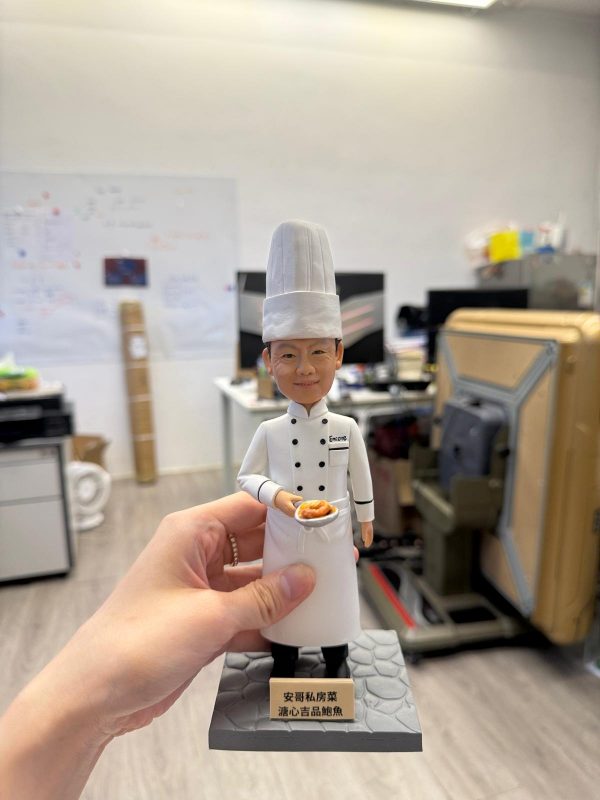 手持展示的主廚公仔：戴高高的白色廚師帽、穿白色雙排扣主廚外套與長圍裙，端著一盤招牌料理站在石紋底座上，前方金色銘牌刻有店名與菜式。