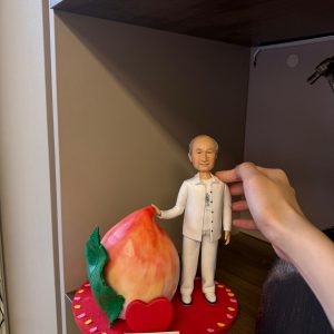 穿白色服飾的長輩人像公仔站在心形紅色底座上，旁邊放著巨型壽桃與祝壽刻字牌