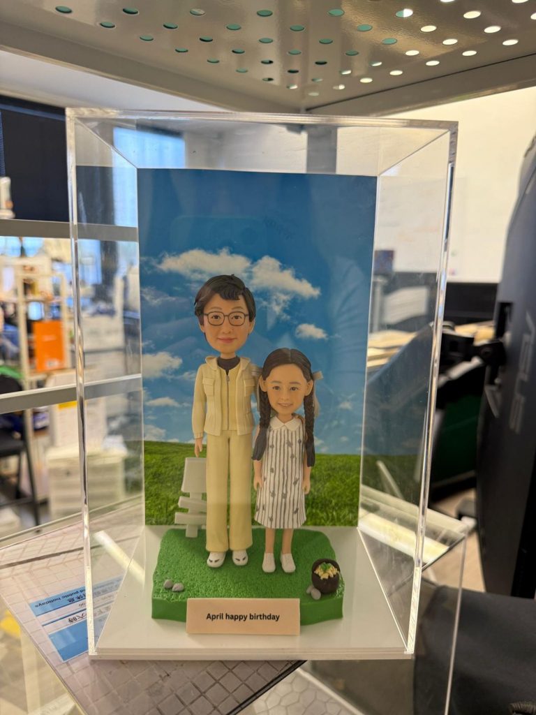 親子雙人公仔站在綠色草地底座上，後方是藍天白雲背板與白色小椅，前方銘牌刻字「April happy birthday」，旁邊擺有小盆栽與石頭點綴
