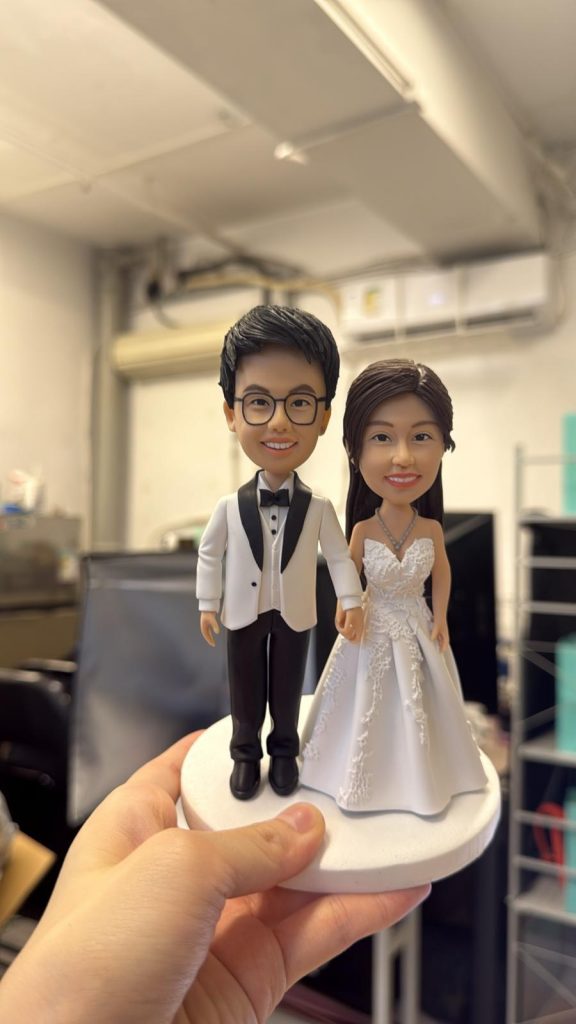 牽手站姿的婚禮公仔：男方戴眼鏡穿白色禮服外套配黑褲與黑色蝴蝶結，女方著蕾絲白紗與項鍊，立於白色圓形底座。