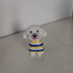 坐姿白色捲毛狗狗公仔，穿黃藍白條紋毛衣，黑鼻圓眼、粉紅舌頭微吐，放置在淺色桌面上。