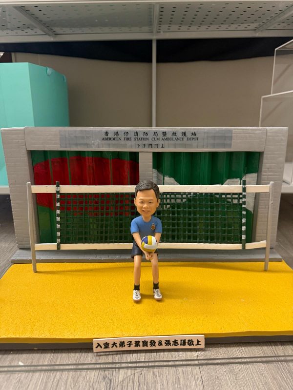 公仔坐在黃綠相間的排球門前，手抱排球，身穿藍色上衣與黑色短褲；背板為「香港仔消防局暨救護站」車房捲門，前方有致敬銘牌。