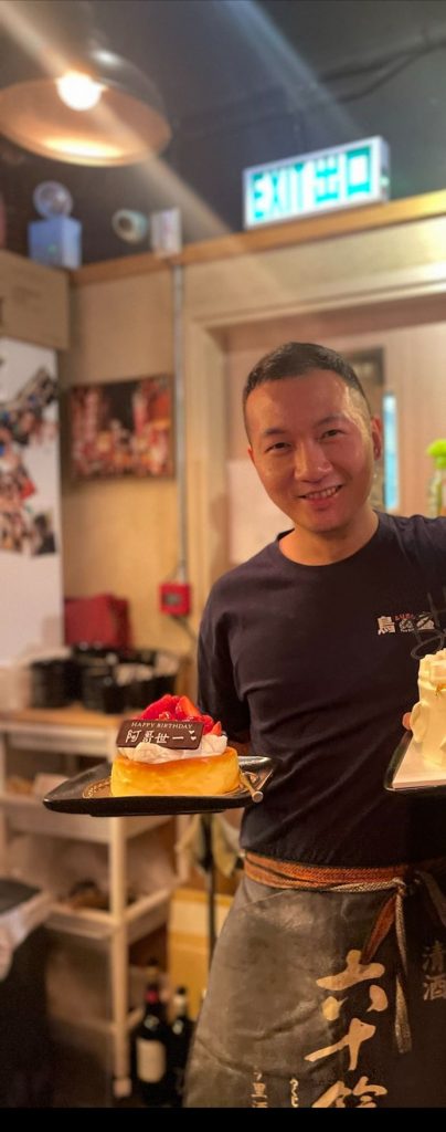 一位身穿黑色T恤和圍裙的男士，笑容滿面地捧著一個士多啤梨生日蛋糕，蛋糕牌寫著「Happy Birthday 阿哥世一」，展現了對職人精神的微縮致敬。