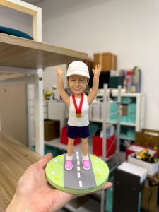 Alt: 女跑者公仔戴白帽掛金牌，雙手高舉站立於公路造型底座，馬拉松完賽紀念客製