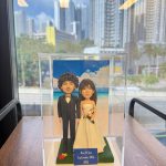 透明展示盒中的婚禮公仔：新郎著黑色西裝與蝴蝶結，新娘穿無肩白紗手捧粉橘花束，腳下綠草地台，背板為藍天白雲與海岸；前方小牌寫著「Keri & Tom September 20th, 2025」。