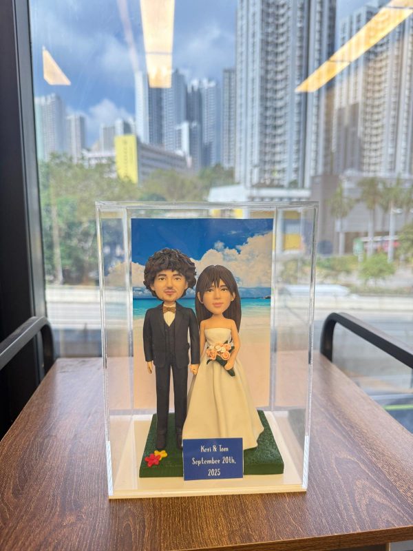 透明展示盒中的婚禮公仔：新郎著黑色西裝與蝴蝶結，新娘穿無肩白紗手捧粉橘花束，腳下綠草地台，背板為藍天白雲與海岸；前方小牌寫著「Keri & Tom September 20th, 2025」。