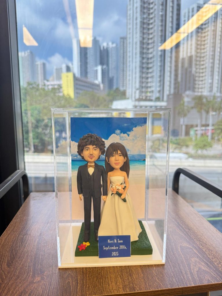 透明展示盒中的婚禮公仔：新郎著黑色西裝與蝴蝶結，新娘穿無肩白紗手捧粉橘花束，腳下綠草地台，背板為藍天白雲與海岸；前方小牌寫著「Keri & Tom September 20th, 2025」。