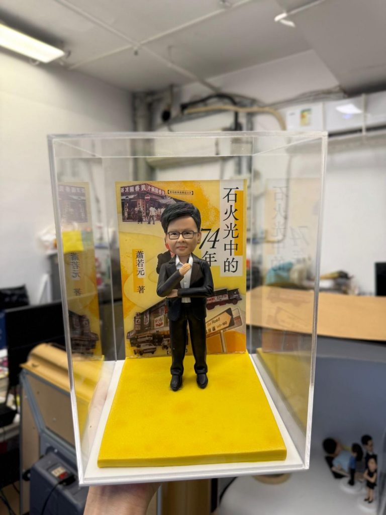 身穿西裝的手工軟陶單人公仔，置於透明展示盒內，背景為黃色書封場景，展現精緻雕塑與立體收藏效果。