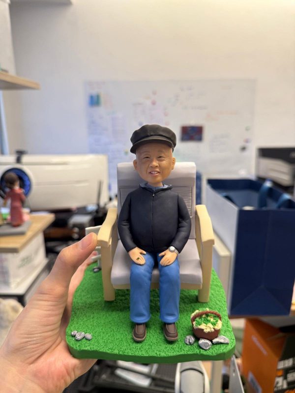 長者人像公仔戴報童帽、穿深色外套與藍褲，坐在木扶手椅上，置於綠色草地底座，旁邊有裝著蔬菜的小籃與石頭點綴，神情和藹