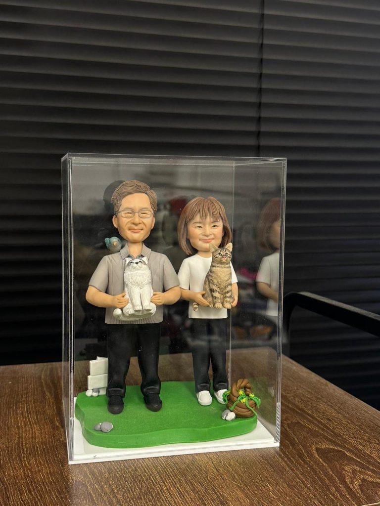 全手工製作的雙人軟陶公仔與一隻貓咪造型，置於透明展示盒與草地場景中，展現細緻神情與溫馨互動。