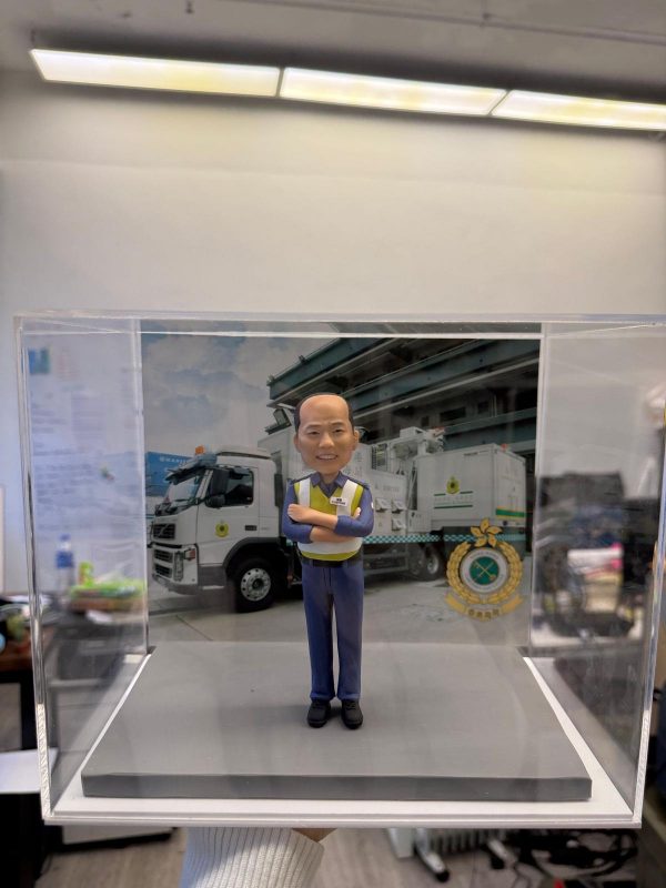 透明展示盒內的人像公仔：光頭男子穿藍色工作褲與反光安全背心，雙手抱臂站在灰色地台上；背板為工程車與作業場景，右下角有圓形徽章標誌。