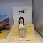 擺設上的女主管公仔：黑長髮、雙手交叉的女士穿米色套裝與荷葉邊襯衫，站在淺木石紋講壇前；前方銘牌寫有「Dilys Cheng, Happy Retirement! Heartfelt thanks for being our Great Leader!」，背景可見 pwc 標誌。