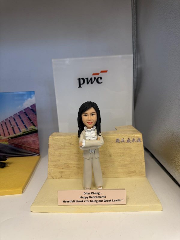 擺設上的女主管公仔：黑長髮、雙手交叉的女士穿米色套裝與荷葉邊襯衫，站在淺木石紋講壇前；前方銘牌寫有「Dilys Cheng, Happy Retirement! Heartfelt thanks for being our Great Leader!」，背景可見 pwc 標誌。