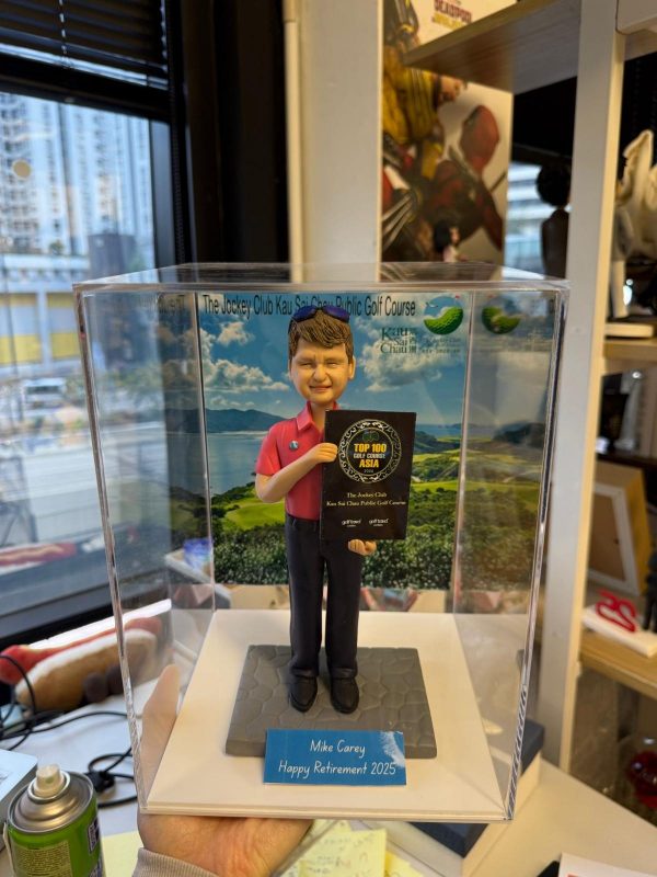 男士人像公仔穿紅色高爾夫上衣與黑長褲，手持「TOP 100 GOLF COURSE ASIA」獎牌站在六角磚地面上，背後是高爾夫球場風景背板，上有「The Jockey Club Kau Sai Chau Public Golf Course」字樣，前方藍色銘牌寫著 “Mike Carey Happy Retirement 2025”