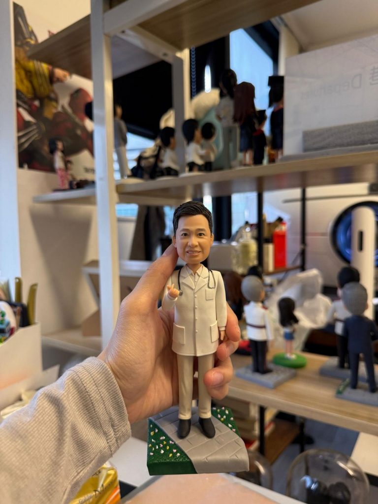 醫師主題人像公仔身穿白袍搭配聽診器，微笑比手勢站在灰色步道與綠地相間的小底座上