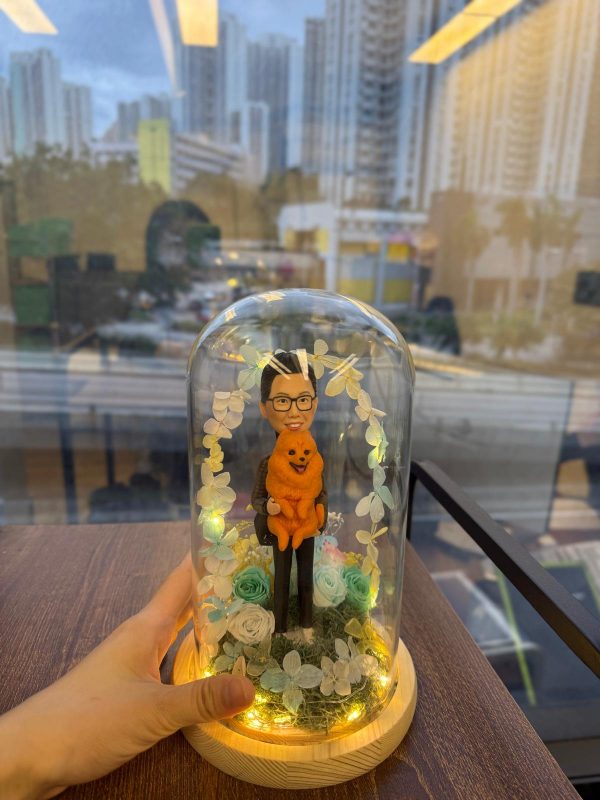 玻璃罩景觀公仔：男子抱著橘色毛茸茸小狗站在苔蘚草地上，周圍點綴藍白永生花與葉片形成拱門，底座為原木色並繞有暖黃燈串發光。