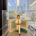 透明展示盒內的制服公仔：短髮男子穿白色襯衫與長褲、黑色皮帶與領帶，做雙手豎拇指手勢站在灰色磚道地台上；身後為綠白相間的病房指示牆與標誌，地台兩側有草皮花帶，前方金色銘牌寫有榮休祝福與科室名稱。